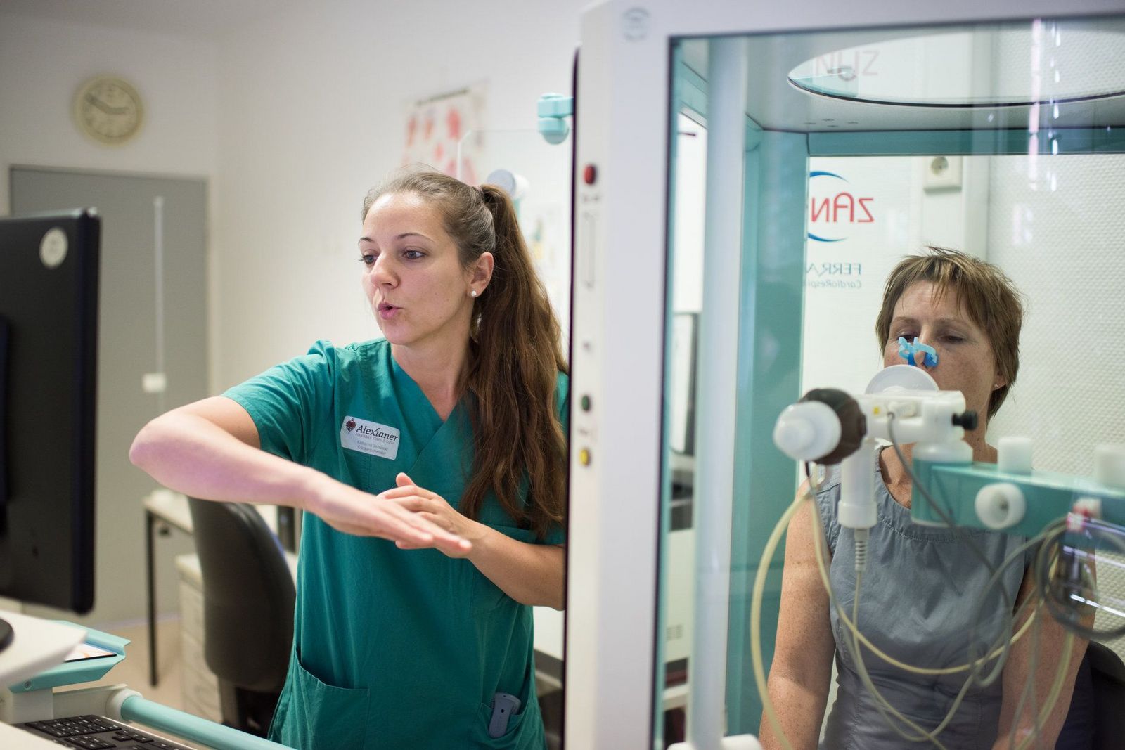 Medizinisch-technische Assistentin führt Lungenfunktionsmessung, die sogenannte Spirometrie, in einer luftdicht abgeschlossenen Glaskabine (Bodyplethysmographie) durch. Die Patientin bläst dabei in ein vorbereitetes Mundstück