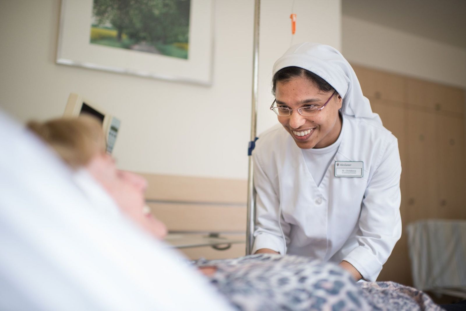 Sympathisch lächelnde Krankenschwester in Ordenstracht beugt sich zu einer älteren Patientin, die im Bett liegt, und spricht mit ihr