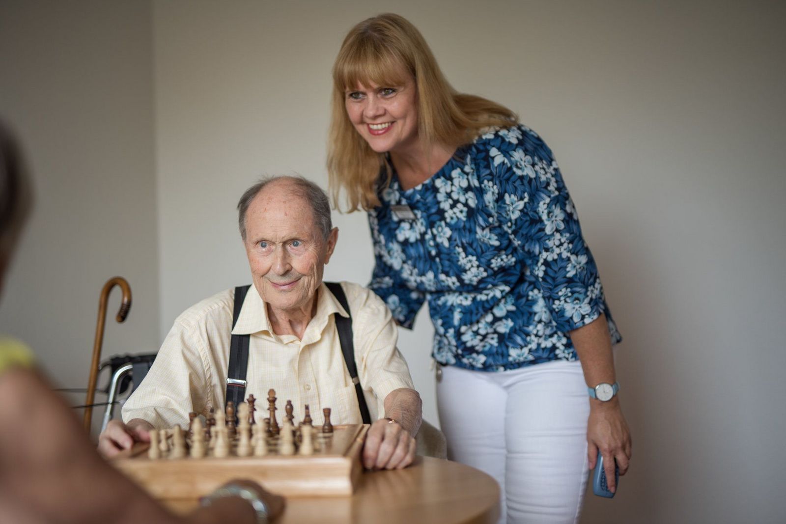 Älterer Herr und ältere Dame spielen Schach. Freundlich zugewandte Krankenschwester steht begleitend zur Seite 